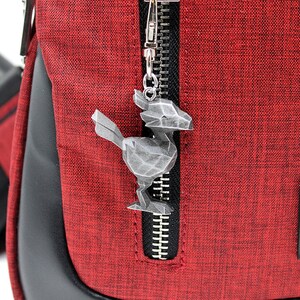 May include: A red fabric messenger bag with a black zipper and a silver metal bird charm attached to the zipper pull. The bag has a black leather trim and a black strap. The bag has a black patch with the text "Where is the heart to Receive Goods where TUOWAN" on it.