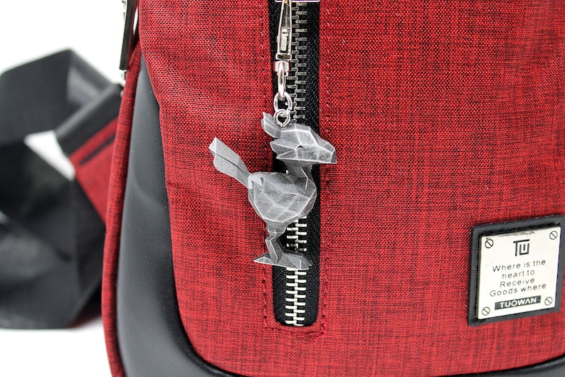 May include: A red fabric messenger bag with a black zipper and a silver metal bird charm attached to the zipper pull. The bag has a black leather trim and a black strap. The bag has a black patch with the text "Where is the heart to Receive Goods where TUOWAN" on it.