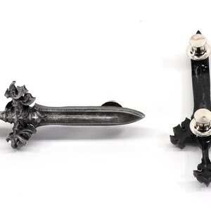 May include: A silver-toned sword pin with intricate details. The pin has a black backing and a silver-toned pin back.