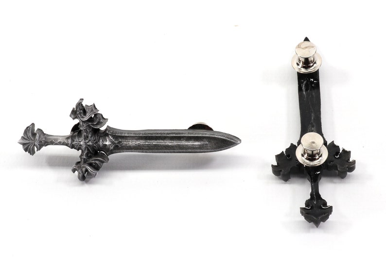 May include: A silver-toned sword pin with intricate details. The pin has a black backing and a silver-toned pin back.