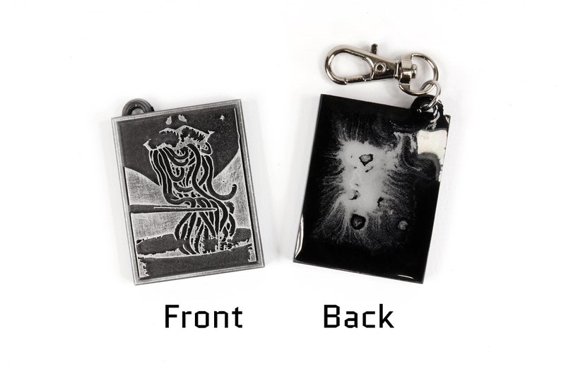 May include: A silver-colored keychain with a black and white image of a creature on the front and a black and white abstract design on the back.