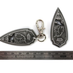 May include: Two silver-colored keychains shaped like a pointed shield with a stylized design of a creature with wings and a crown. The keychains are attached to a silver-colored metal ring and a silver-colored metal clip.