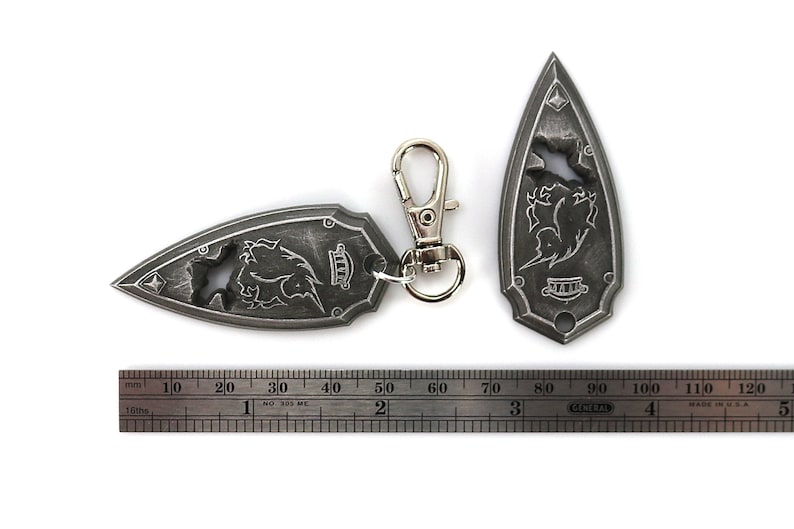May include: Two silver-colored keychains shaped like a pointed shield with a stylized design of a creature with wings and a crown. The keychains are attached to a silver-colored metal ring and a silver-colored metal clip.
