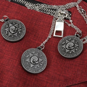 May include: Three silver-tone pendant necklaces with circular, engraved pendants featuring a star design and text. The necklaces rest on a textured red fabric, near a silver zipper.
