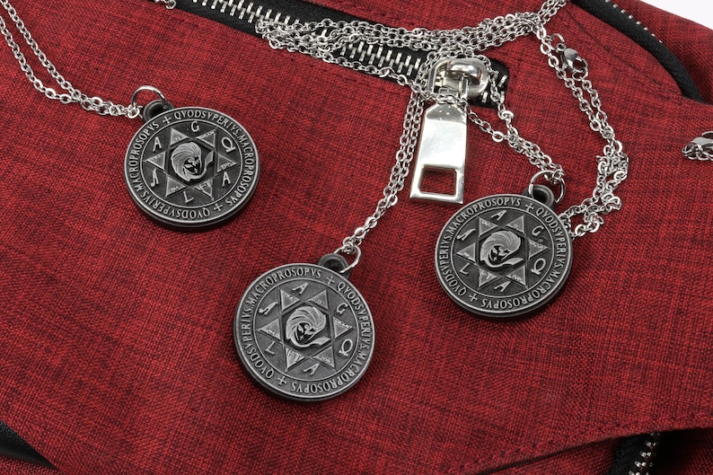 May include: Three silver-tone pendant necklaces with circular, engraved pendants featuring a star design and text. The necklaces rest on a textured red fabric, near a silver zipper.