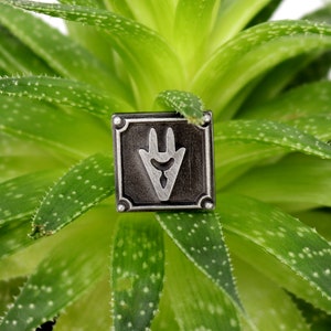Op de afbeelding: Een zilverkleurige metalen pin met een zwarte achtergrond en een gestileerd handontwerp. De pin zit op een groene vetplant.