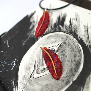 May include: Two red feather pendants with gold accents on a black background. The feathers are on a black cord necklace.