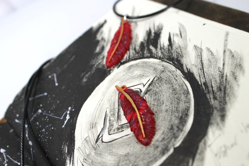May include: Two red feather pendants with gold accents on a black background. The feathers are on a black cord necklace.