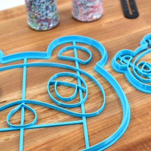 May include: Two blue plastic cookie cutters shaped like animal faces. The larger cutter is a detailed outline of a raccoon face. The smaller cutter is a simpler outline of a panda face.