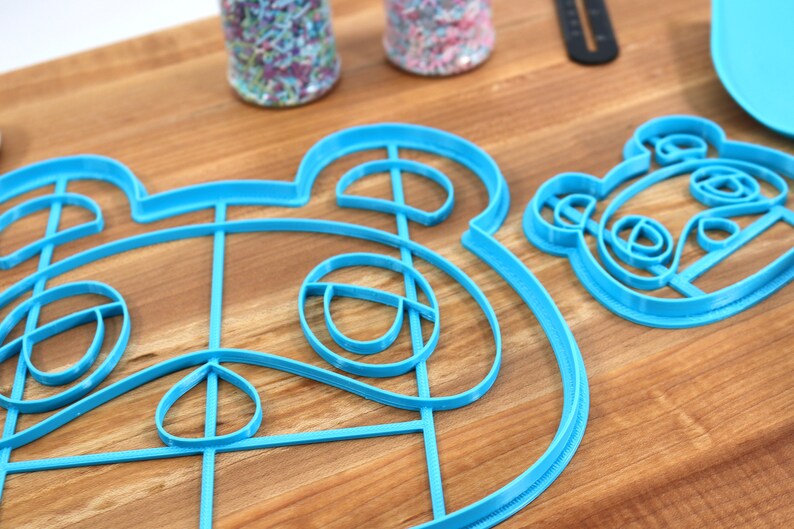 May include: Two blue plastic cookie cutters shaped like animal faces. The larger cutter is a detailed outline of a raccoon face. The smaller cutter is a simpler outline of a panda face.