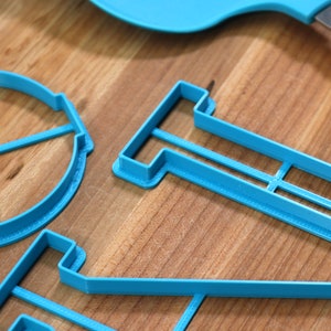 May include: A set of three blue plastic cookie cutters shaped like a circle with four lines, a rectangle with a line across the middle, and a funnel. The cutters are on a wooden surface.