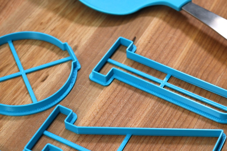 May include: A set of three blue plastic cookie cutters shaped like a circle with four lines, a rectangle with a line across the middle, and a funnel. The cutters are on a wooden surface.
