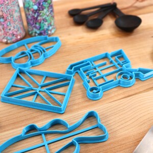 May include: A set of blue plastic cookie cutters in various shapes, including a fish, a diamond, a house, a bus, and a pair of glasses. The cutters are on a wooden surface.