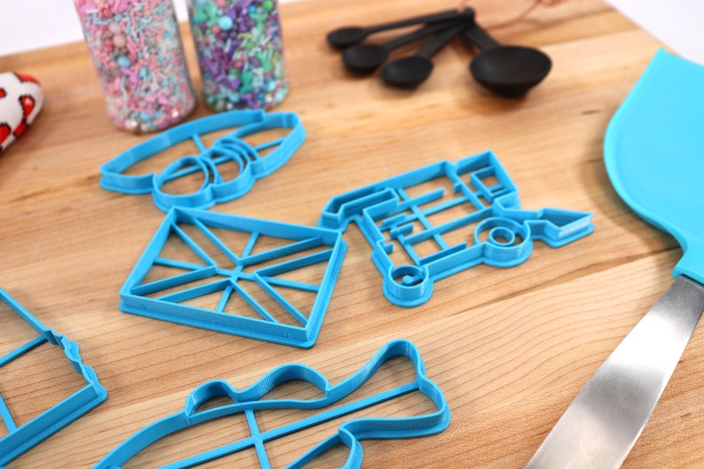 May include: A set of blue plastic cookie cutters in various shapes, including a fish, a diamond, a house, a bus, and a pair of glasses. The cutters are on a wooden surface.