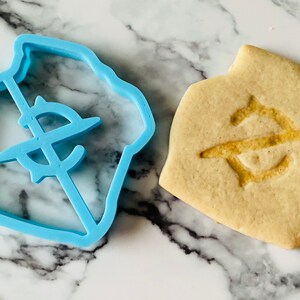 May include: A blue plastic cookie cutter shaped like a house with a dollar sign inside. Next to it is a baked cookie with the same design.