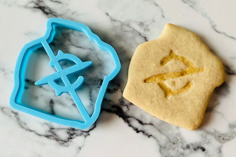May include: A blue plastic cookie cutter shaped like a house with a dollar sign inside. Next to it is a baked cookie with the same design.