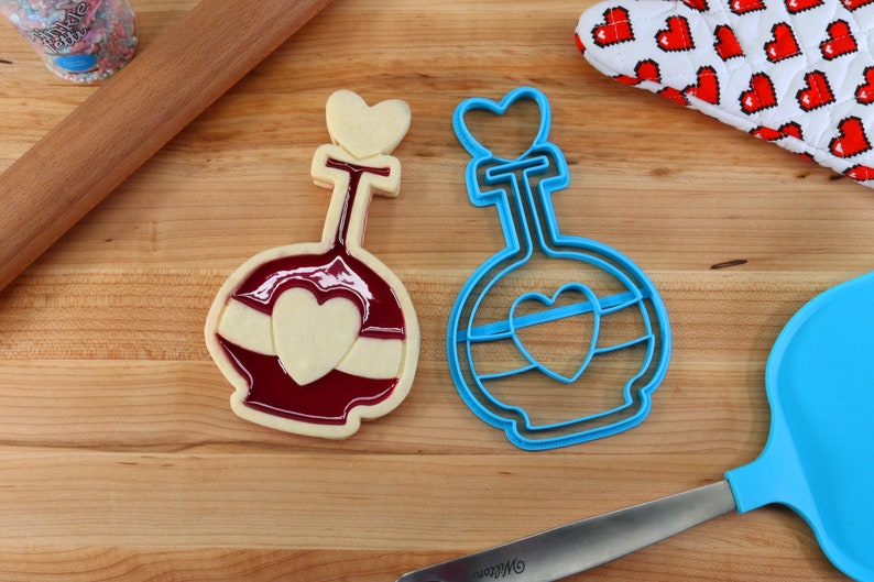 May include: Two cookie cutters in the shape of a bottle with a heart on the front. One cutter is blue and the other is red with white icing.