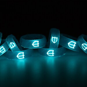 May include: A group of eight glow-in-the-dark silicone rings with a shield logo. The rings are stacked and arranged in a pile. The rings are a bright turquoise color and glow brightly in the dark.