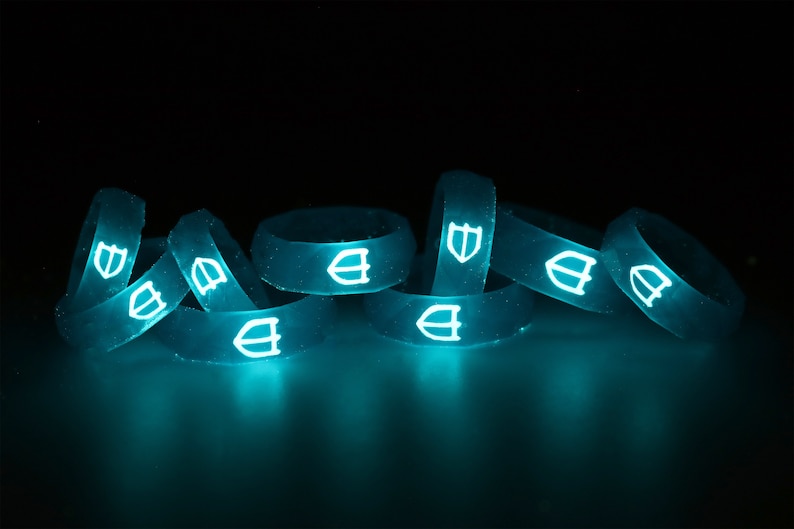 May include: A group of eight glow-in-the-dark silicone rings with a shield logo. The rings are stacked and arranged in a pile. The rings are a bright turquoise color and glow brightly in the dark.