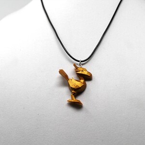 May include: A gold-colored pendant necklace featuring a geometric, stylized bird design. The pendant is attached to a black cord.