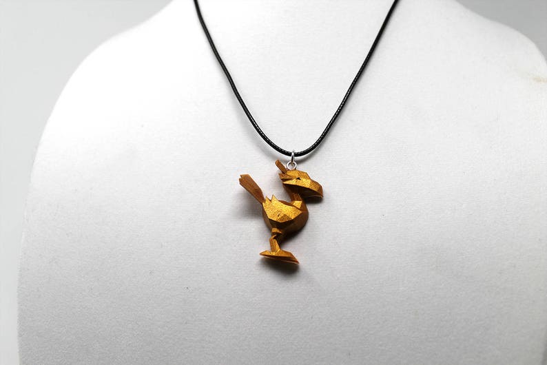 May include: A gold-colored pendant necklace featuring a geometric, stylized bird design. The pendant is attached to a black cord.