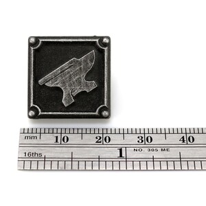 May include: A silver metal pin with a black background and a silver anvil design. The pin is 2 cm wide.