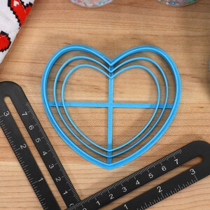May include: A blue plastic heart-shaped cookie cutter with three concentric rings. The cutter is on a wooden surface with a black ruler.