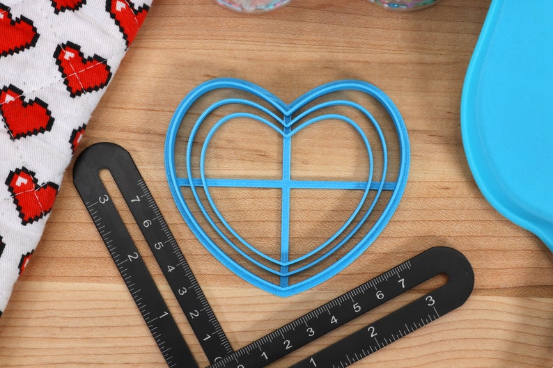 May include: A blue plastic heart-shaped cookie cutter with three concentric rings. The cutter is on a wooden surface with a black ruler.