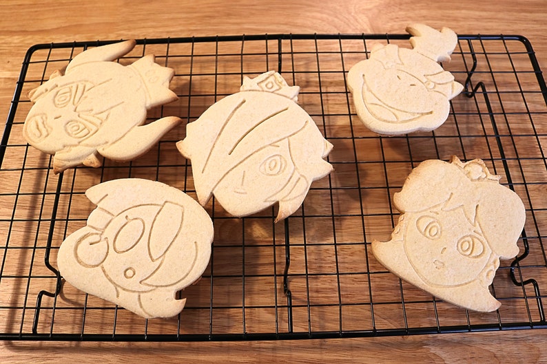 May include: Five unbaked sugar cookies on a wire rack. The cookies are shaped like cartoon faces and have been decorated with a simple outline.