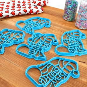 May include: Five blue plastic Christmas cookie cutters shaped like Santa Claus. The cutters are on a wooden surface.