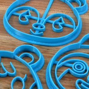 May include: Three blue plastic cookie cutters shaped like cartoon animal faces. The cutters are on a wooden surface.