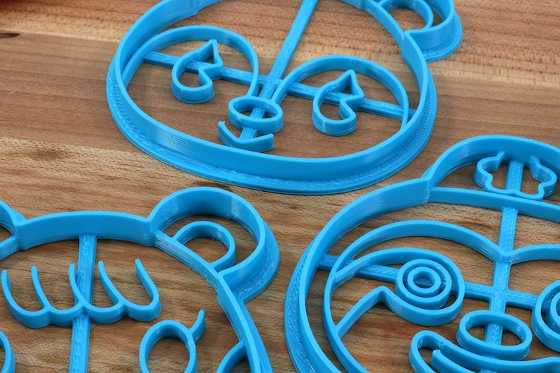 May include: Three blue plastic cookie cutters shaped like cartoon animal faces. The cutters are on a wooden surface.