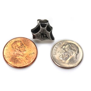 May include: A silver-colored metal earring with a five-pointed star shape. The star has a textured surface and a black kanji character in the center. The earring is shown next to two US coins, a penny and a dime, for size comparison.
