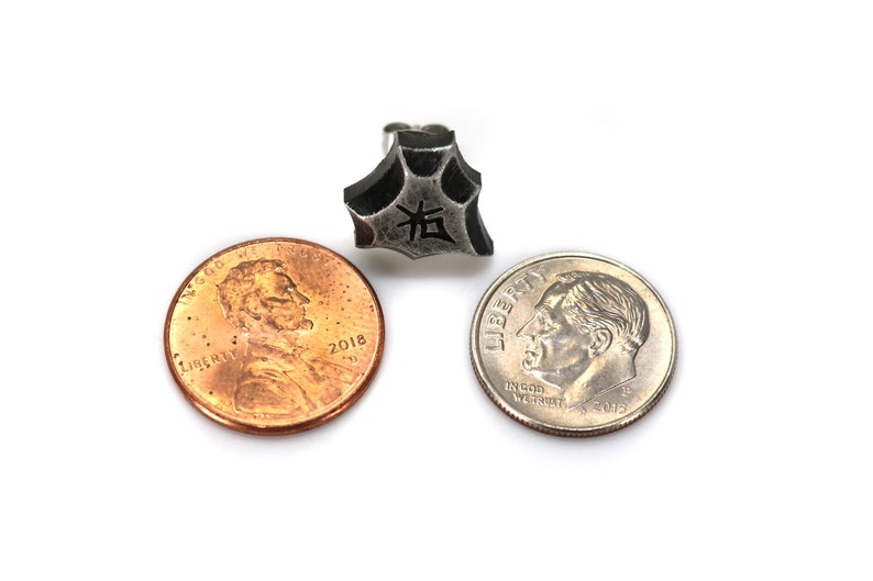 May include: A silver-colored metal earring with a five-pointed star shape. The star has a textured surface and a black kanji character in the center. The earring is shown next to two US coins, a penny and a dime, for size comparison.