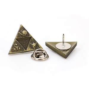 May include: A gold-toned triangular pin with a design of a stylized triangle with four symbols inside. The back of the pin has a silver-toned pin back.