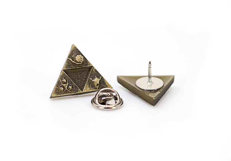 May include: A gold-toned triangular pin with a design of a stylized triangle with four symbols inside. The back of the pin has a silver-toned pin back.
