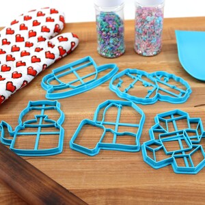 May include: A set of blue plastic cookie cutters shaped like a teapot, a cup, a house, a jar, and a stack of cubes. The cutters are on a wooden cutting board with a red and white oven mitt, a rolling pin, and two jars of sprinkles.