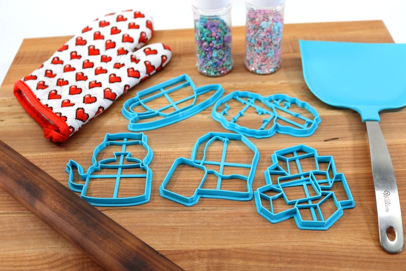 May include: A set of blue plastic cookie cutters shaped like a teapot, a cup, a house, a jar, and a stack of cubes. The cutters are on a wooden cutting board with a red and white oven mitt, a rolling pin, and two jars of sprinkles.