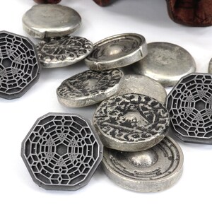May include: A collection of silver-toned metal coins with intricate designs. Some coins feature a geometric pattern, while others have a more traditional design. The coins are scattered on a white surface.