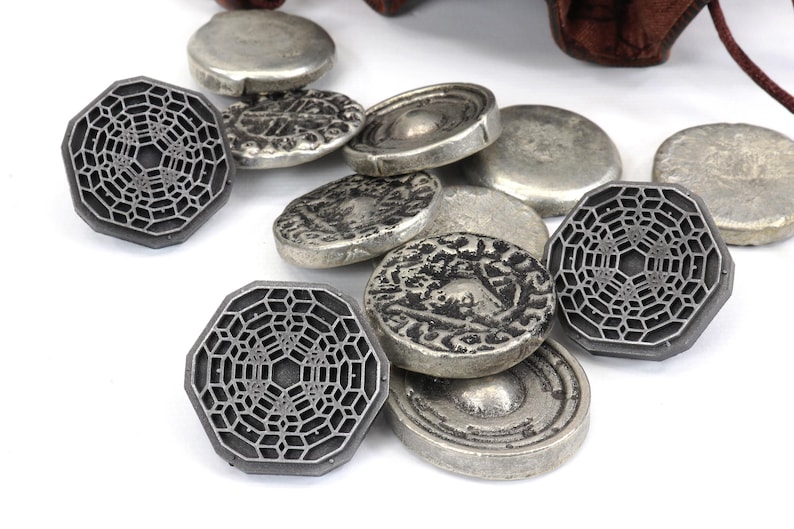 May include: A collection of silver-toned metal coins with intricate designs. Some coins feature a geometric pattern, while others have a more traditional design. The coins are scattered on a white surface.