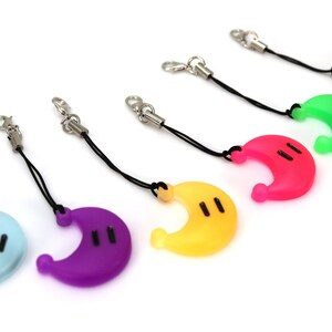 May include: A set of seven colorful crescent moon-shaped phone charms with black eyes. Each charm is attached to a black string with a silver clip.