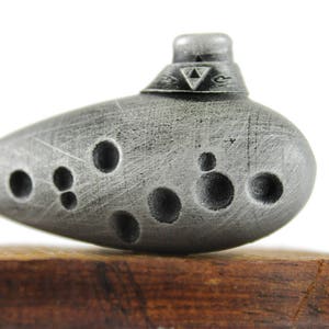 Puede incluir: Una ocarina plateada con un s&iacute;mbolo triangular en la parte superior. La ocarina tiene varios agujeros y una superficie texturizada.