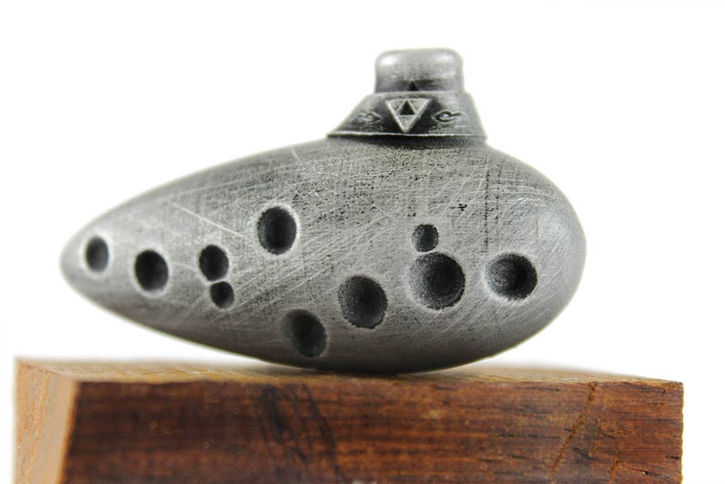 Puede incluir: Una ocarina plateada con un s&iacute;mbolo triangular en la parte superior. La ocarina tiene varios agujeros y una superficie texturizada.