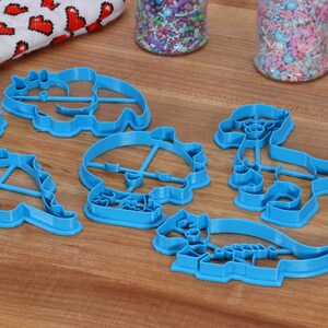 May include: A set of six blue plastic dinosaur-shaped cookie cutters on a wooden surface.