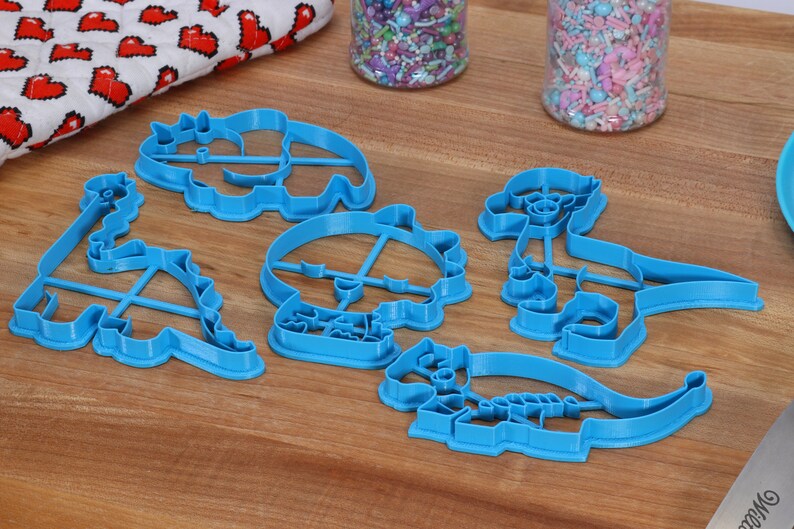 May include: A set of six blue plastic dinosaur-shaped cookie cutters on a wooden surface.