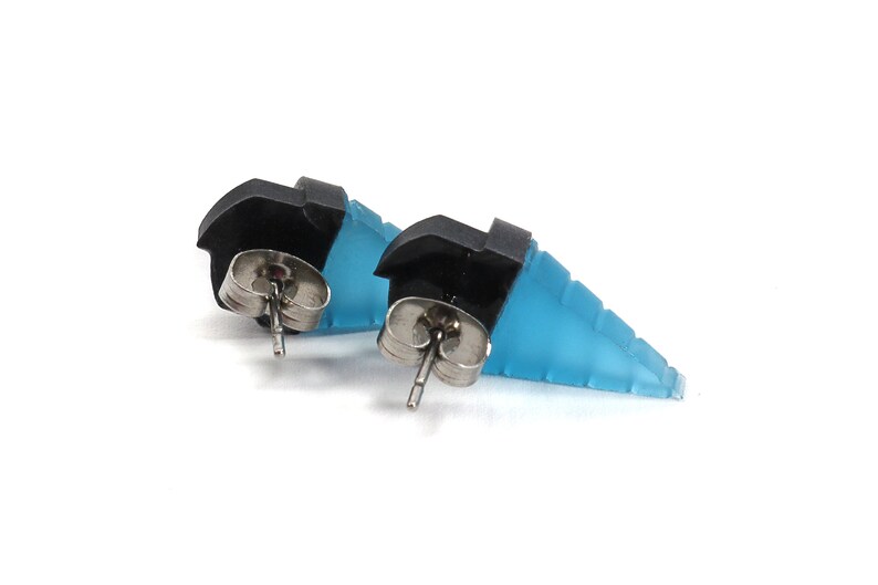May include: A pair of stud earrings featuring a geometric design. The earrings have a black top section and a light blue, stepped lower section. The earring posts are silver-colored.