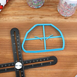 May include: A blue plastic cookie cutter shaped like a house with four windows. The cutter is on a wooden surface with a black ruler.