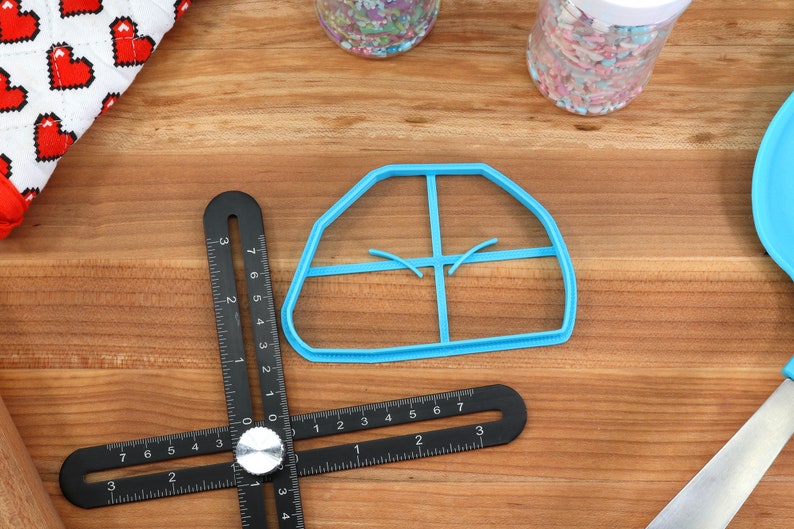 May include: A blue plastic cookie cutter shaped like a house with four windows. The cutter is on a wooden surface with a black ruler.
