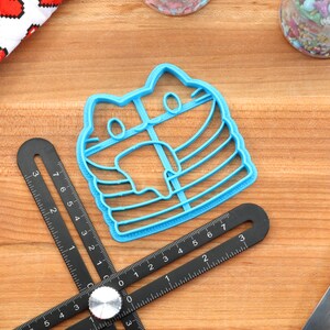 May include: A blue plastic cookie cutter shaped like a cat wearing a striped mask. The cutter is on a wooden surface with a ruler and a rolling pin.