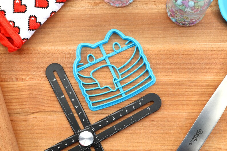 May include: A blue plastic cookie cutter shaped like a cat wearing a striped mask. The cutter is on a wooden surface with a ruler and a rolling pin.
